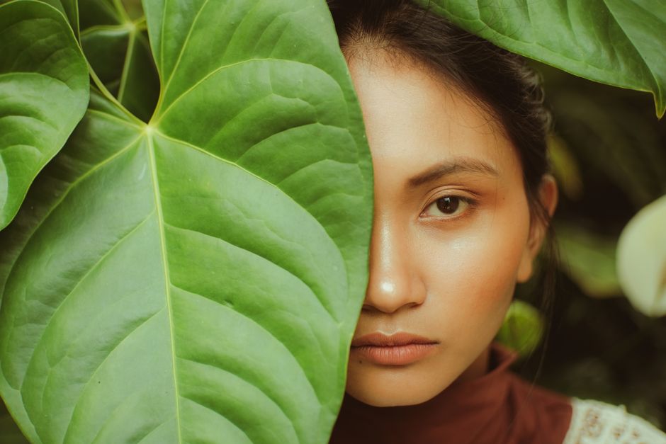 A serene portrait of a woman partially concealed by a large green leaf in a natural setting.