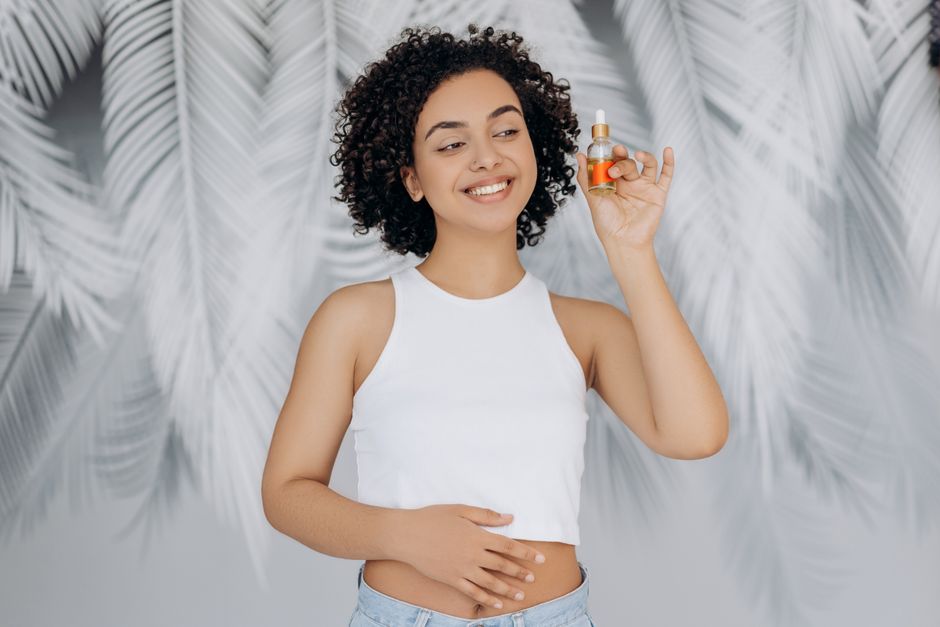 Smiling woman with curly hair holding essential oil bottle in contemporary indoor setting.