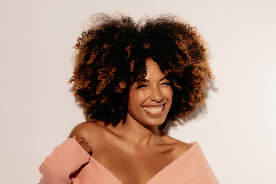 Cheerful woman with curly afro hair in pink attire, smiling brightly against a neutral background.