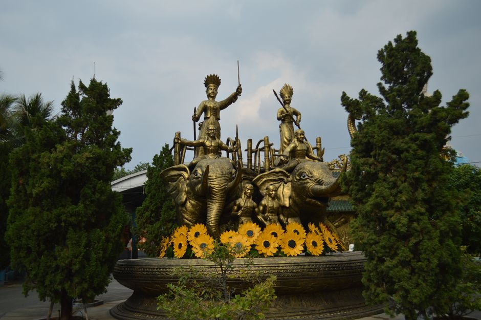 A stunning golden statue featuring elephants and figures, surrounded by greenery in an outdoor temple garden.