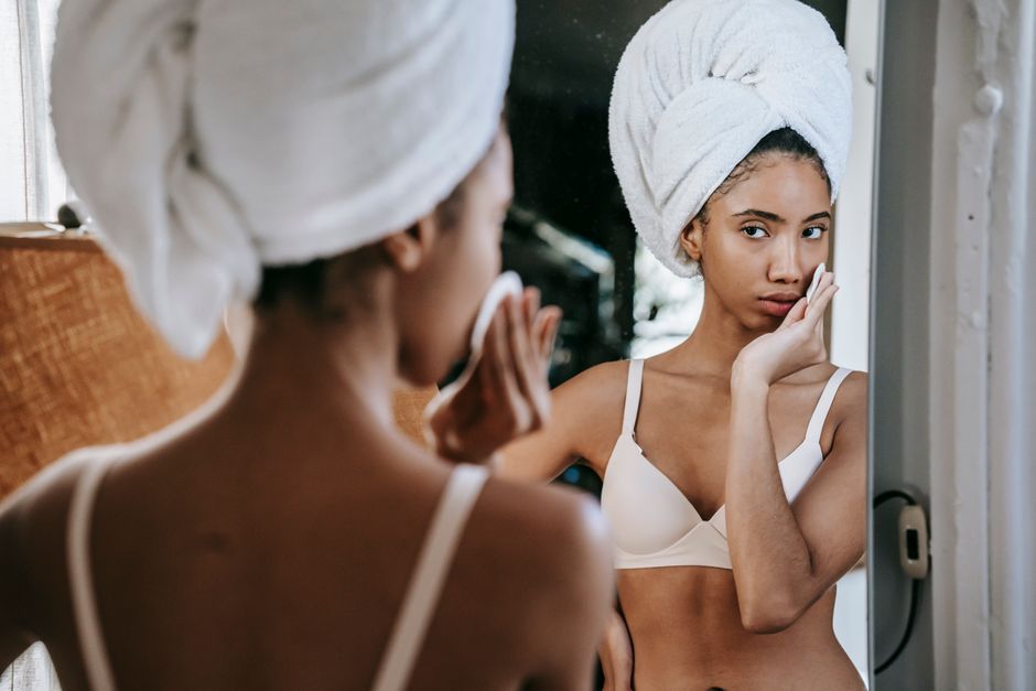Serious ethnic female looking at mirror while moisturizing skin with lotion during daily beauty routine in bathroom