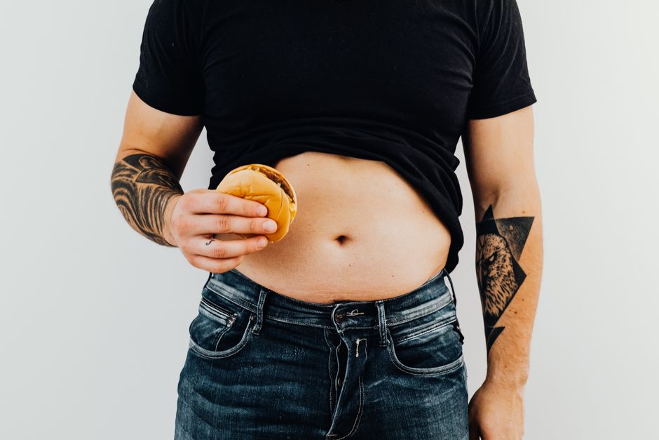 Adult man holding a hamburger, displaying tattoos and abdomen, wearing casual clothing.