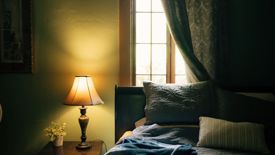 A dimly lit bedroom with a glowing lamp, soft pillows, and elegant curtains by the window.