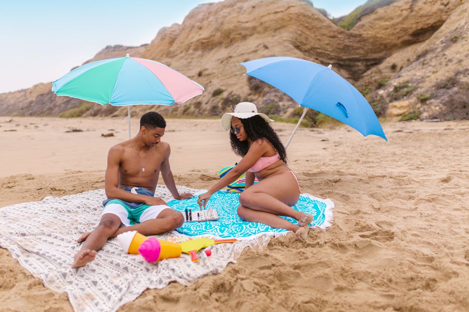 A couple playing chess on a sandy beach under colorful umbrellas during a summer day.