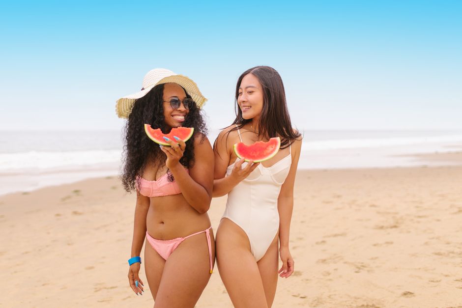 Two women enjoying a sunny day at the beach, relishing juicy watermelon slices. Perfect summer vibes.