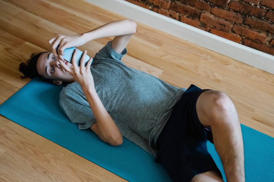 High angle of fit young male in sportswear lying on yoga mat on floor while using mobile phone at home
